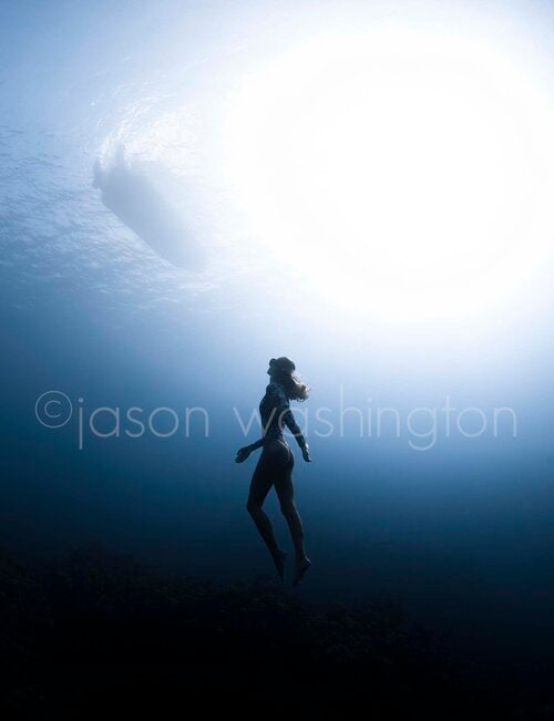Underwater shot looking upward at a freediver rising along a deep vertical wall, silhouetted against a bright surface light with the faint shape of a boat visible overhead.