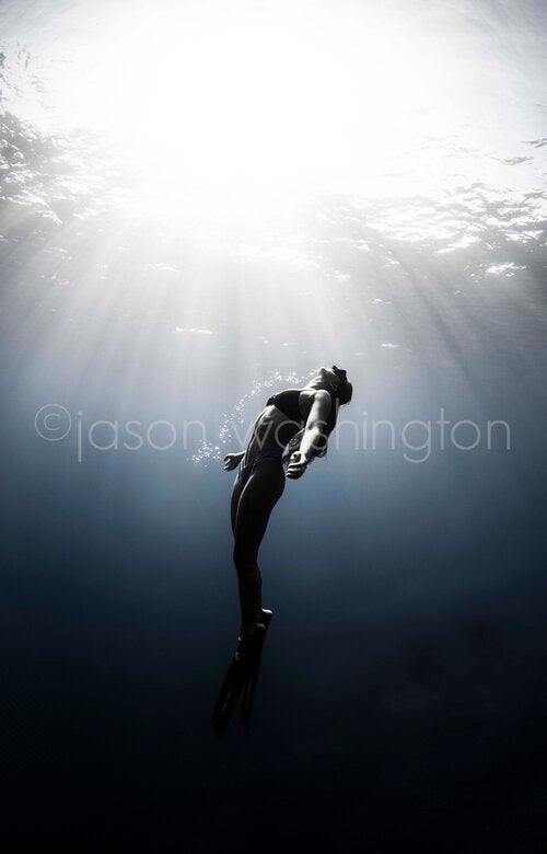 Black-and-white underwater photograph of a freediver ascending toward the bright water surface, arms open and fins trailing, with beams of sunlight streaming down through clear water.