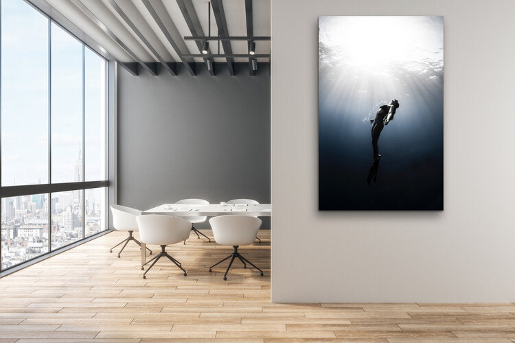 Black-and-white underwater photograph of a freediver ascending toward the bright water surface, arms open and fins trailing, with beams of sunlight streaming down through clear water.