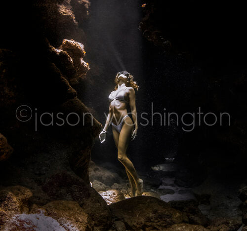 Freediver in a bikini standing on the floor of a dark underwater cave, looking up as a shaft of sunlight breaks through an opening above, illuminating the rocky walls around her
