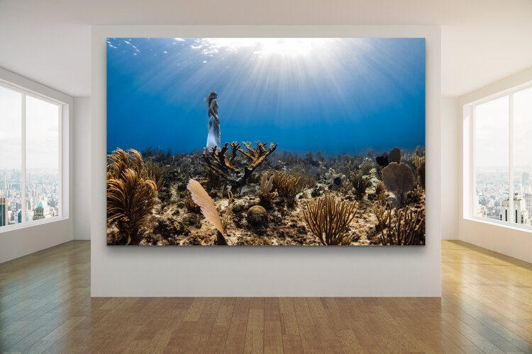 Underwater scene of a woman in a flowing white dress standing on a coral-covered seafloor, bathed in beams of sunlight streaming down through deep blue water.