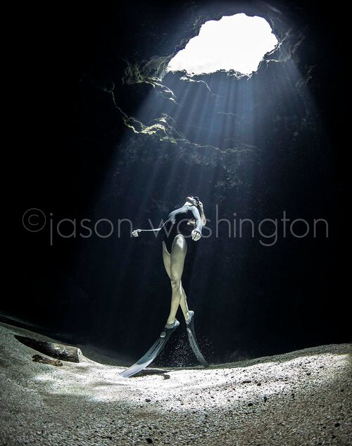 Underwater view of a freediver in a black swimsuit standing on a sandy spring floor beneath a cavern opening, as shafts of sunlight stream down through the clear water into the dark grotto.