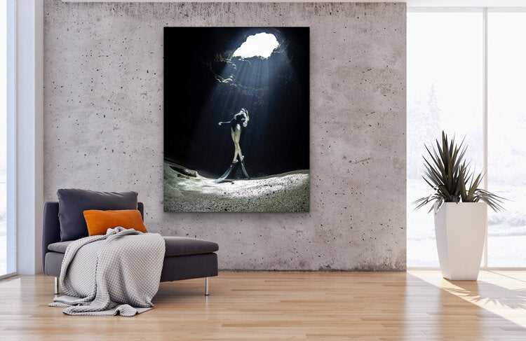 Underwater view of a freediver in a black swimsuit standing on a sandy spring floor beneath a cavern opening, as shafts of sunlight stream down through the clear water into the dark grotto.