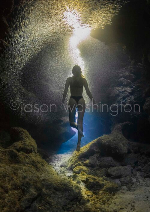 Underwater photograph of a freediver rising through a rocky cavern as a dense school of small silver fish swirls overhead, with a beam of sunlight piercing down from the surface