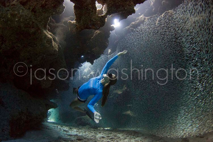 Freediver in a blue wetsuit gliding through a coral cave as a dense school of tiny silvery fish swirls around her, with light beams filtering down from openings above.