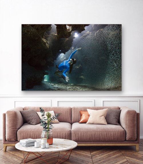 Freediver in a blue wetsuit gliding through a coral cave as a dense school of tiny silvery fish swirls around her, with light beams filtering down from openings above.