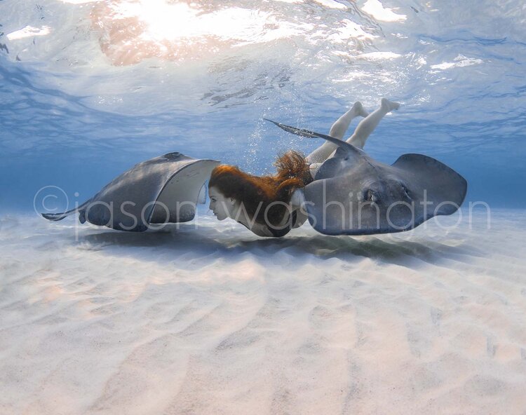 A freediver with red hair swimming between two southern stingrays over rippled white sand in shallow, sunlit waters.