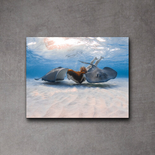 A freediver with red hair swimming between two southern stingrays over rippled white sand in shallow, sunlit waters.