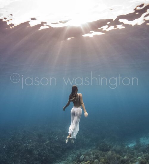 Underwater photograph of a freediver in a flowing white skirt rising above a coral reef as sunbeams stream down through the glassy surface like shafts of rain.