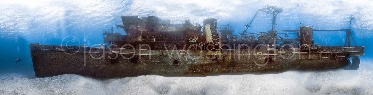 Panoramic underwater photograph of the entire USS Kittiwake submarine rescue shipwreck on the seabed, with a diver beside the bow and sunlight filtering down through blue water.