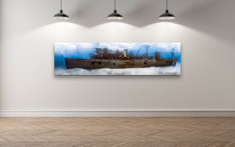 Panoramic underwater photograph of the entire USS Kittiwake submarine rescue shipwreck on the seabed, with a diver beside the bow and sunlight filtering down through blue water.