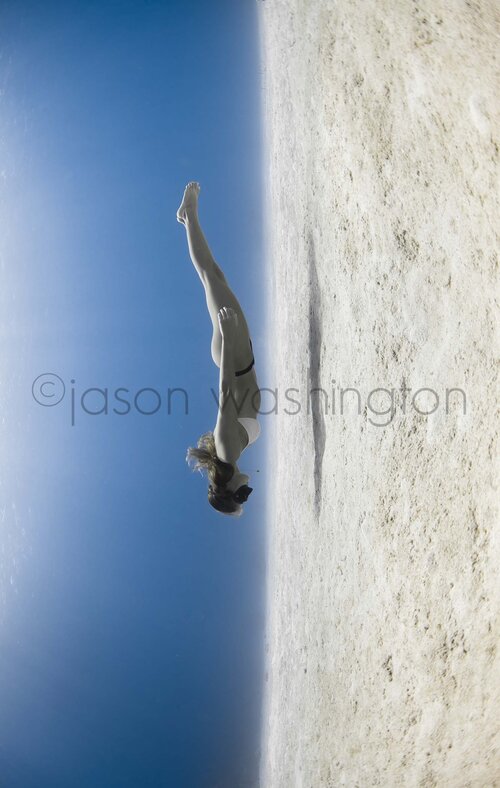 Vertical underwater photograph of a freediver in a white bikini diving headfirst between a glassy blue surface and a sandy seafloor, creating a mirrored, upside-down effect.