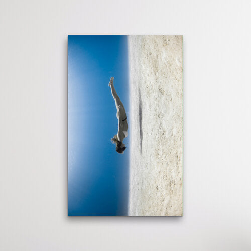 Vertical underwater photograph of a freediver in a white bikini diving headfirst between a glassy blue surface and a sandy seafloor, creating a mirrored, upside-down effect.