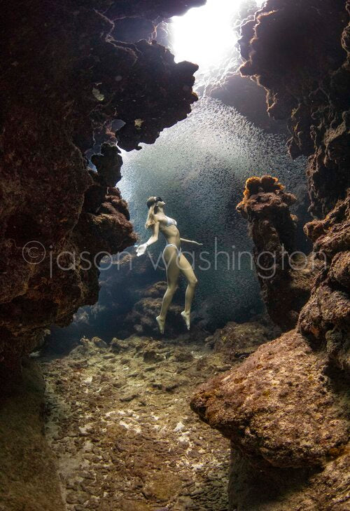 Underwater photograph of a freediver in a white bikini floating between rocky walls of a cavern as a dense school of tiny silver fish swirls above her under a shaft of sunlight.