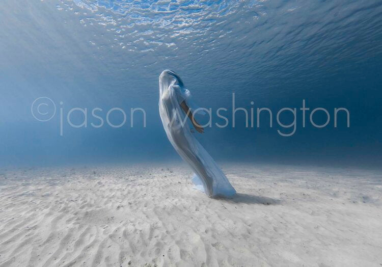 Underwater photograph of a freediver draped in a sheer white fabric, leaning back as she ascends through clear, blue water above a rippled sandy seabed.