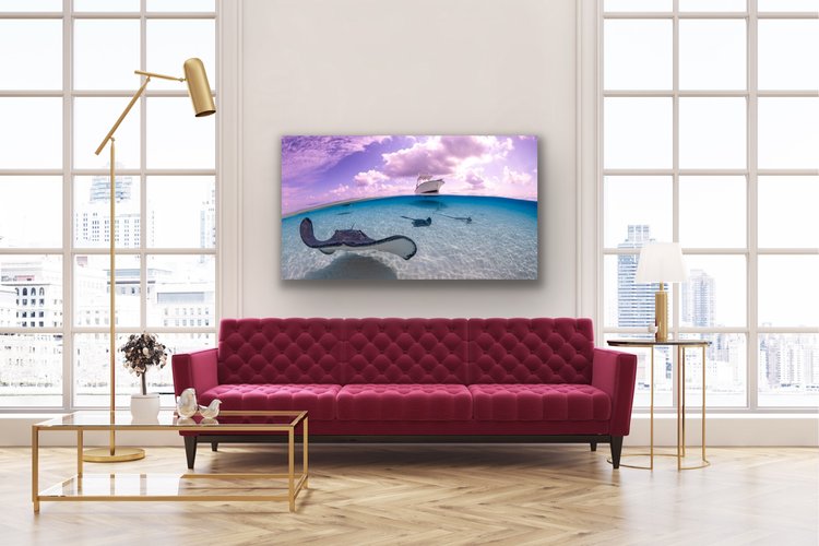 Split-level underwater photograph of a southern stingray gliding over rippled white sand with two other stingrays behind, and a small boat floating on the glassy turquoise sea above under a partly cloudy sky.