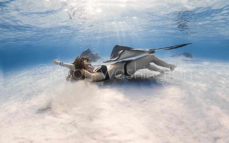 A freediver lying on a sandy sea floor with arms outstretched, surrounded by several southern stingrays gliding overhead in clear, sunlit water.